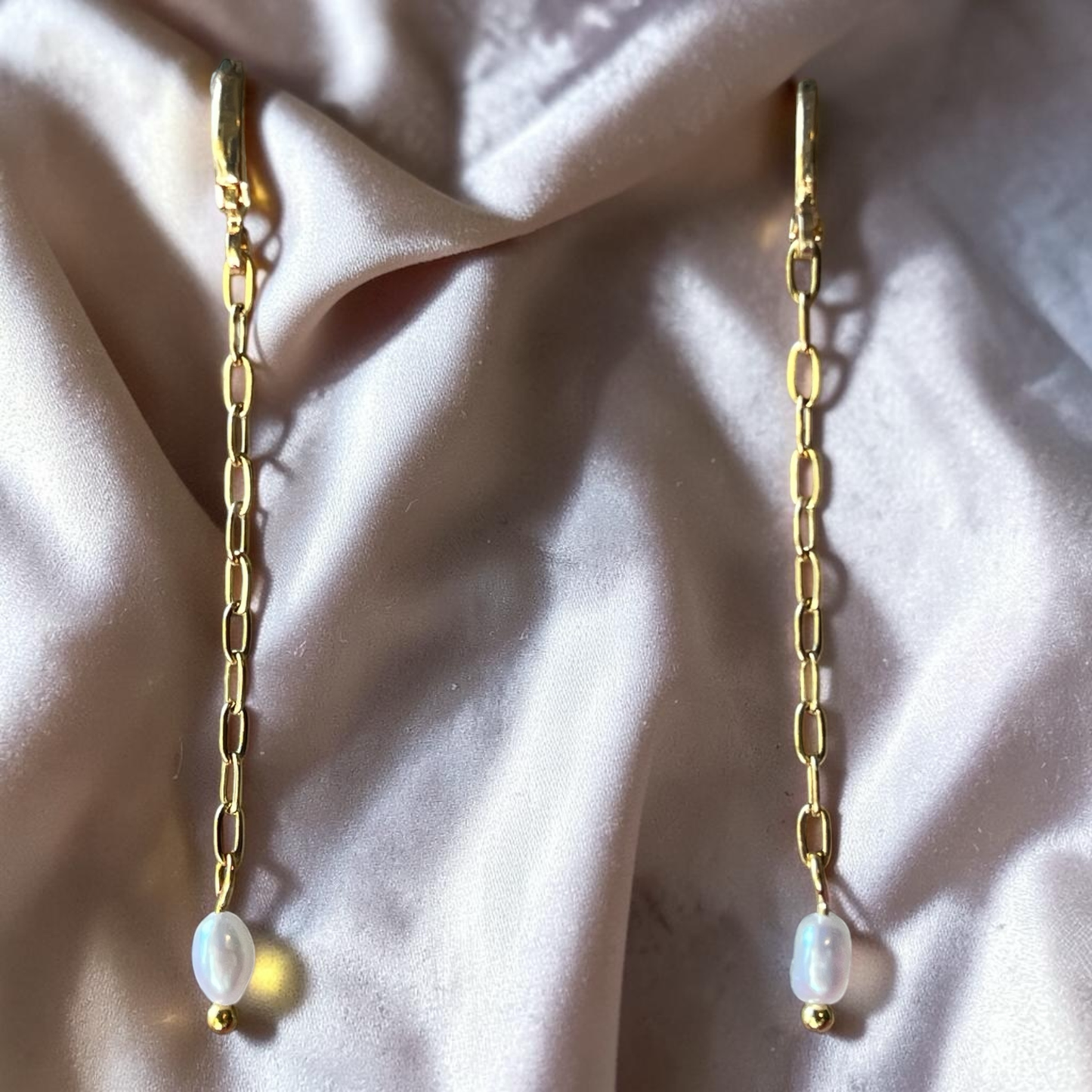 Gold chain earrings with pearl drops on a soft pink fabric background