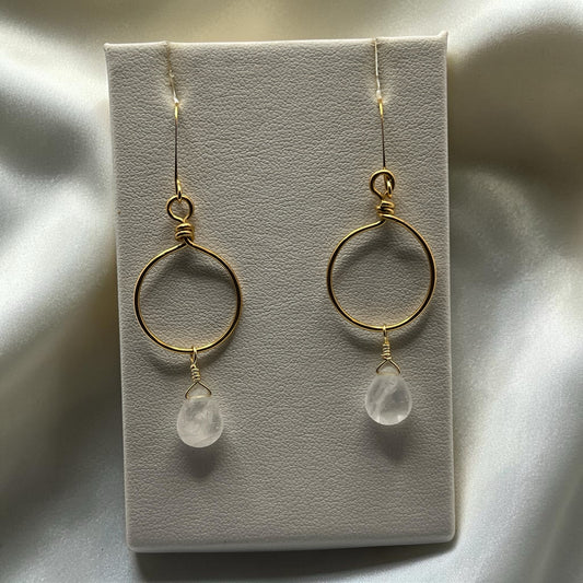 Blakeley Lane Jewelry gold-tone wire-wrapped hoop earrings with teardrop clear crystal accents on a display stand.