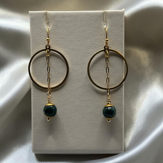 Blakeley Lane Jewelry gold-tone circular hoop earrings with hanging faceted deep green beads on a white display stand.