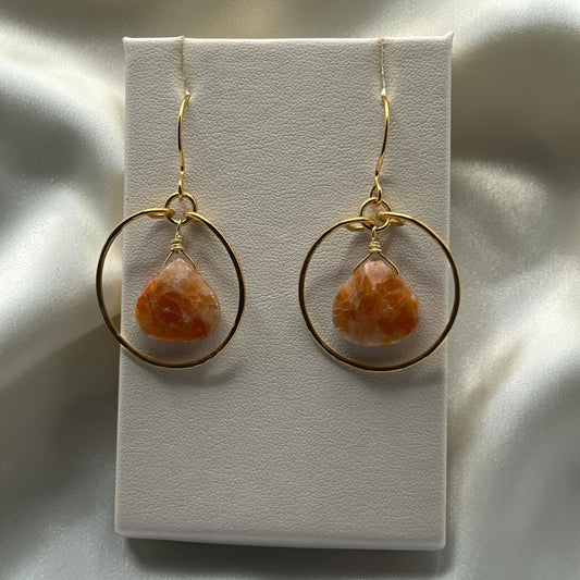 Blakeley Lane Jewelry teardrop orange gemstone earrings featuring polished gold-tone hoops on a white display stand.