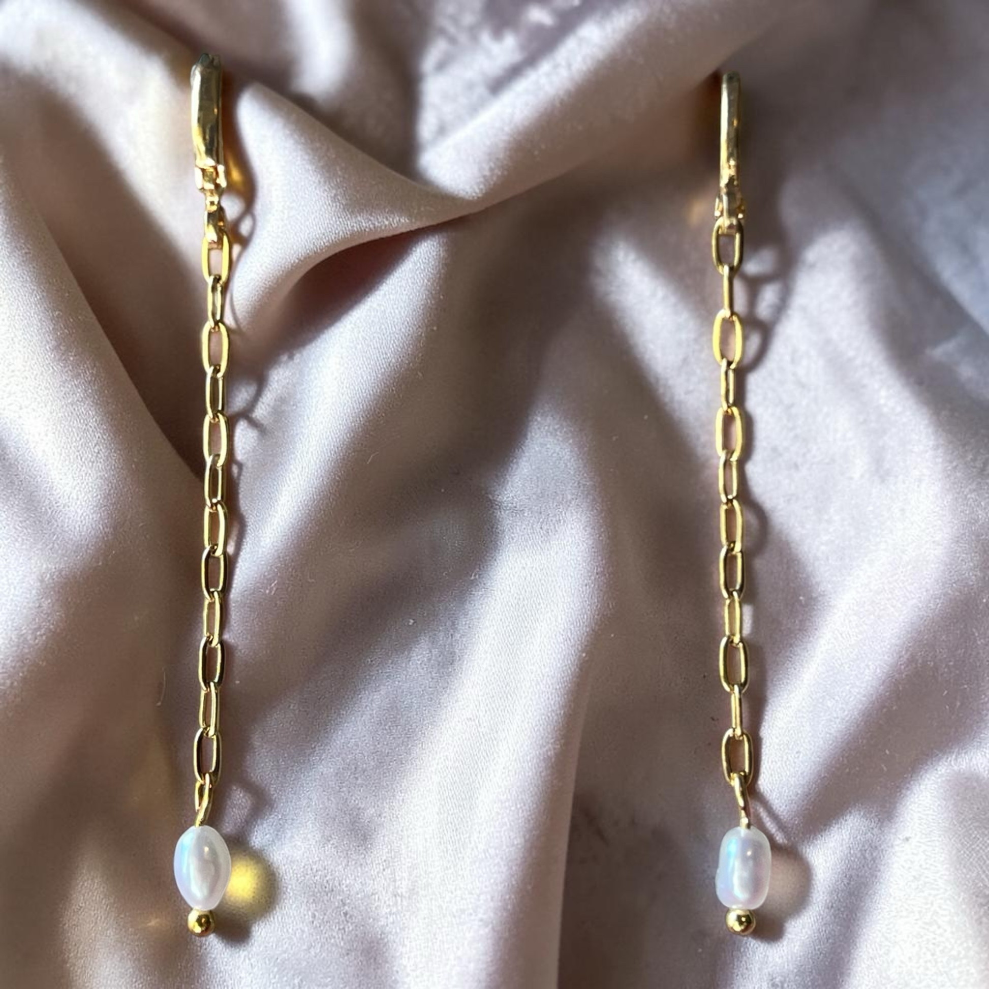 Gold chain earrings with pearl drops on a soft pink fabric background