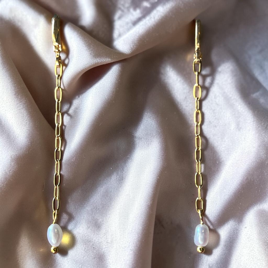 Gold chain earrings with pearl drops on a soft pink fabric background
