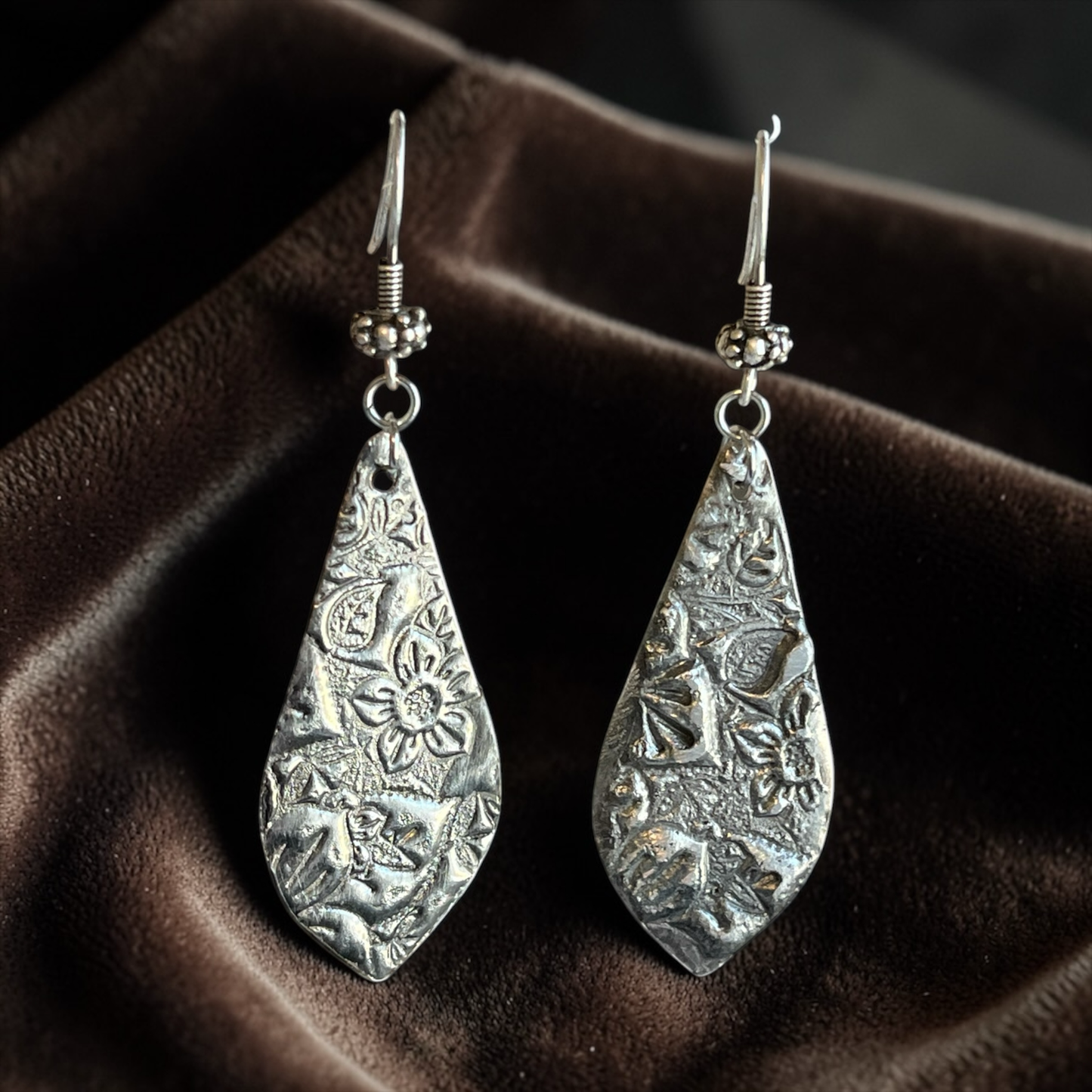 Pair of silver teardrop earrings with intricate patterns on a brown fabric background