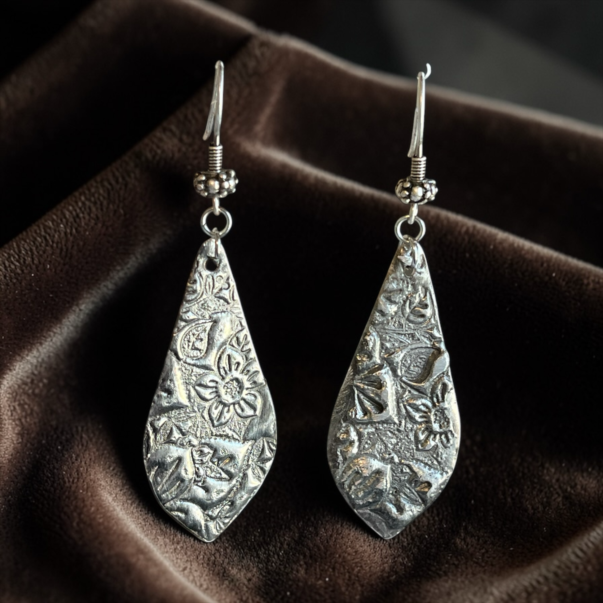 Pair of silver teardrop earrings with intricate patterns on a brown fabric background